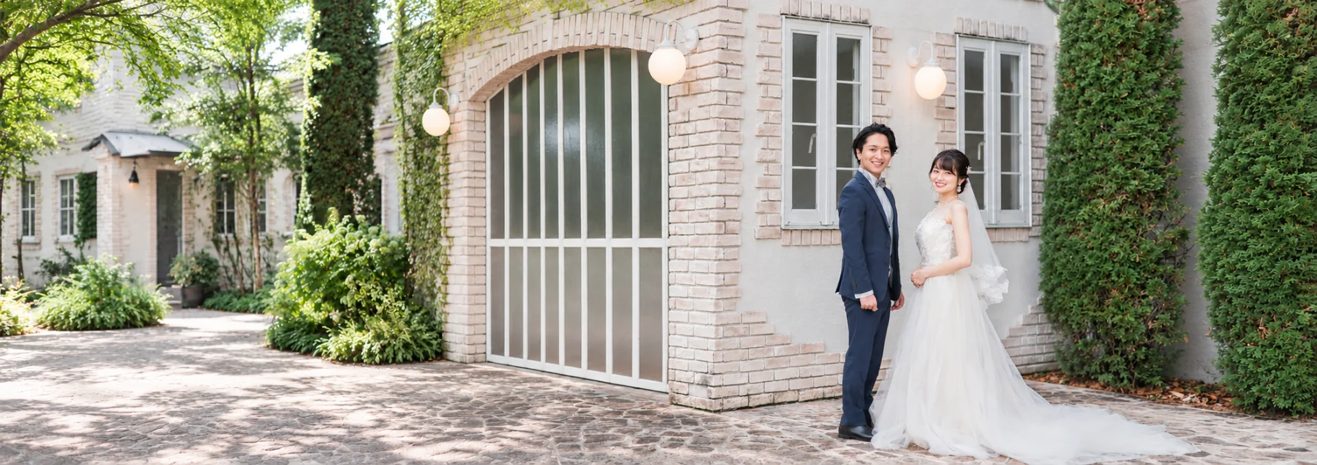 緑に囲まれたレンガ造りの洋館の前でこちらを向いて微笑むウェディング姿の男女