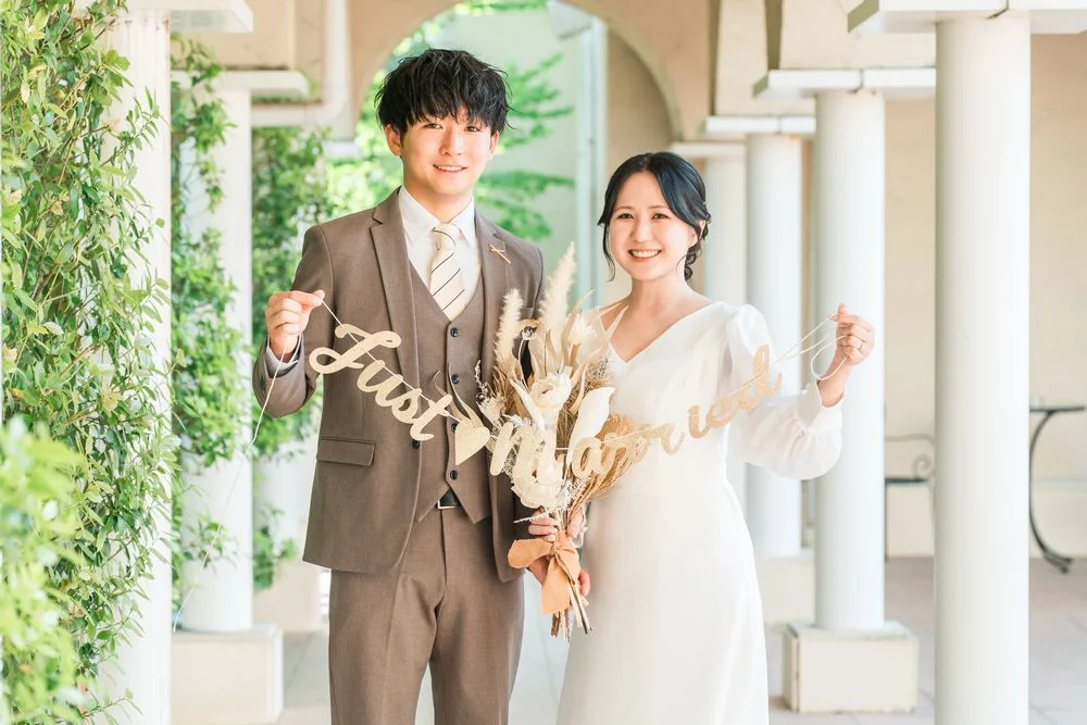 緑に囲まれた回廊で「Just Married」のサインを手に微笑むウェディング姿の男女